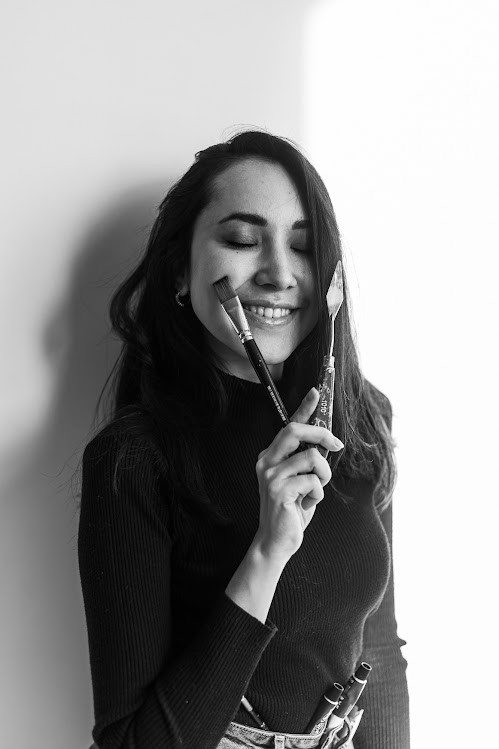 Black-and-white portrait of Aliya, eyes closed and smiling, holding a paintbrush and a palette knife