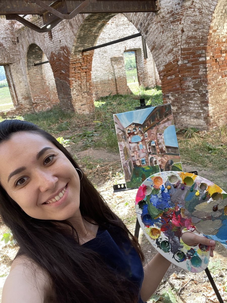 Aliya with her palette in front of an easel among old brick arches