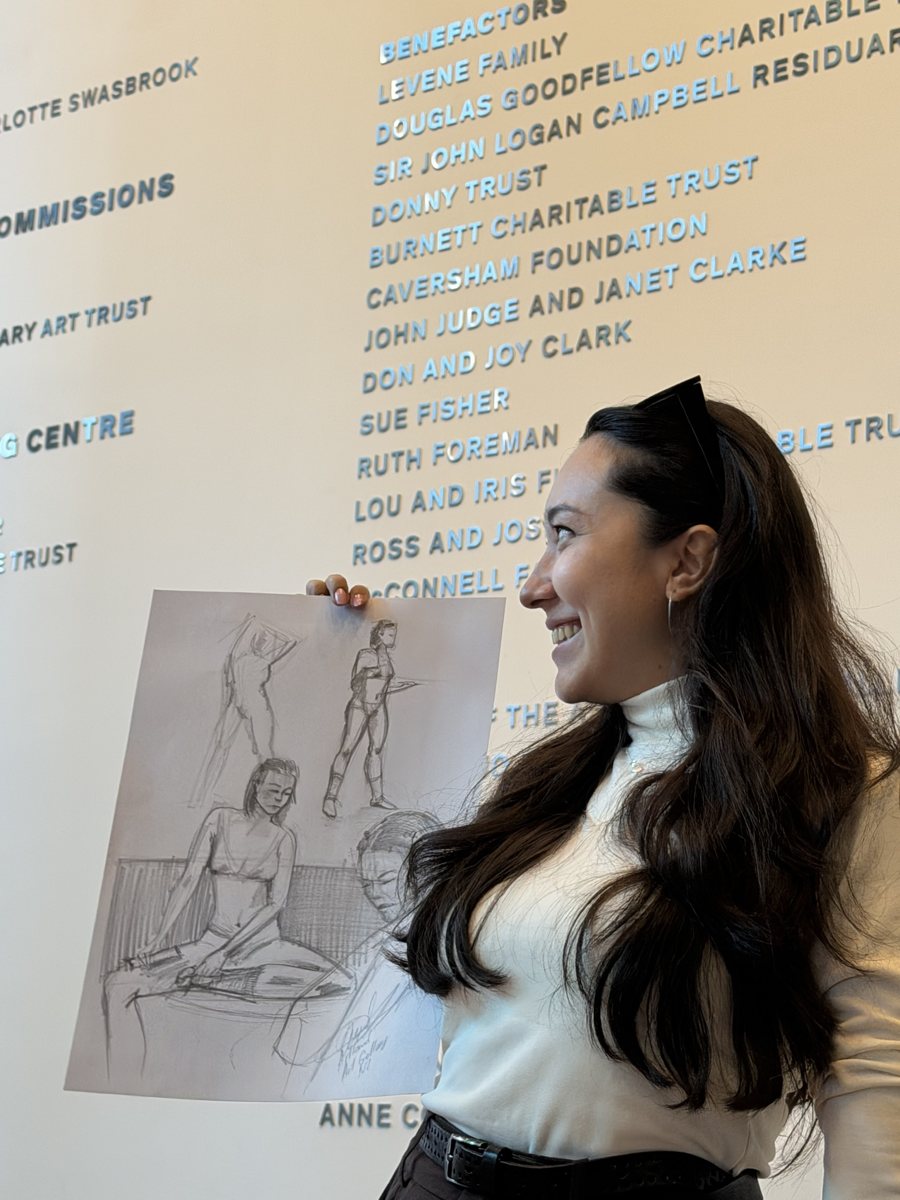Aliya smiling as she holds up a sheet of figure sketches in an art gallery
