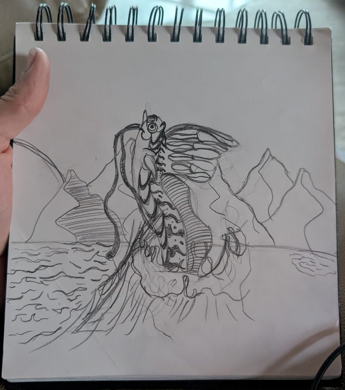 Kyle's pencil drawing in a spiral sketchbook: a winged fantasy creature rising over mountains and the sea