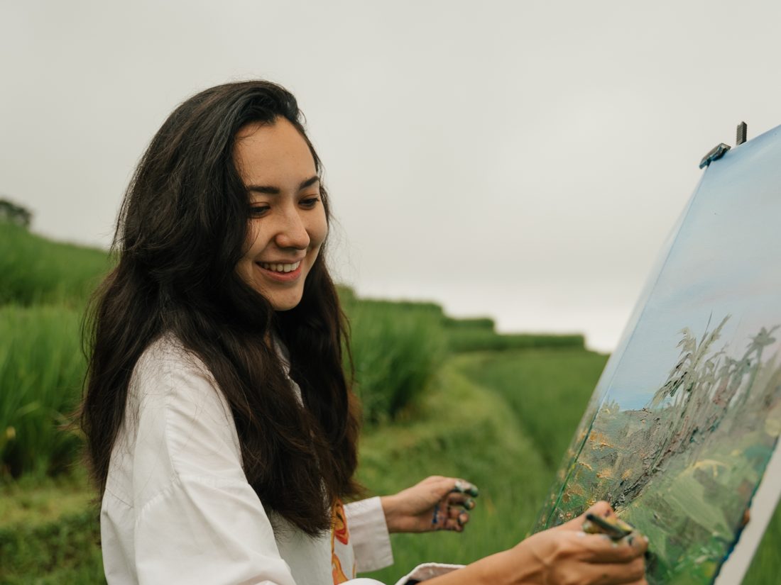 Aliya smiling as she paints a landscape at an easel among Bali's rice fields
