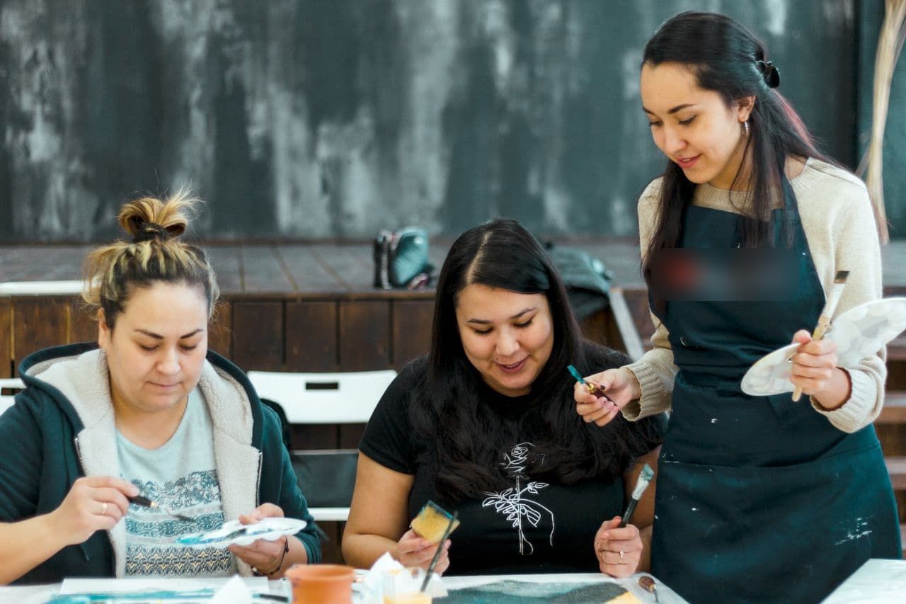 Aliya showing two students a brush technique at the painting table