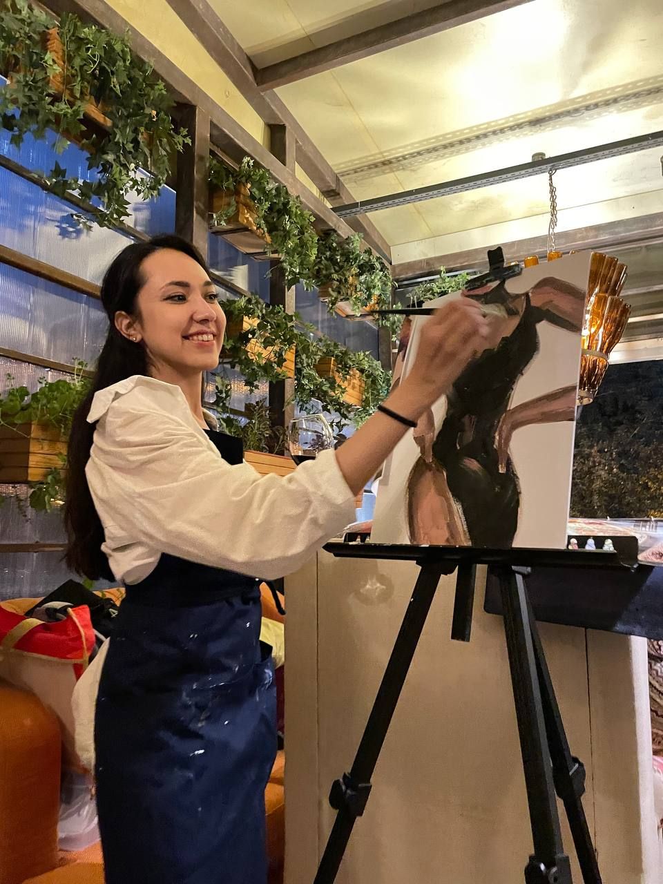 Aliya smiling while painting a figure on canvas at an evening live-painting event