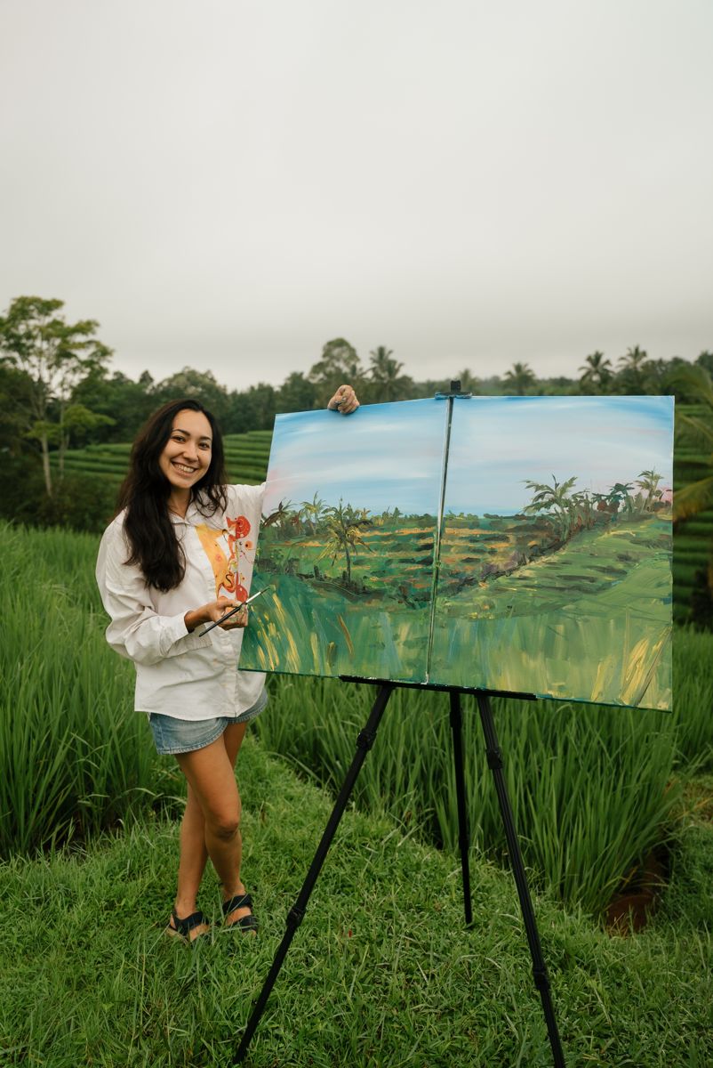 Aliya smiling next to her finished diptych of rice terraces on an easel in Bali