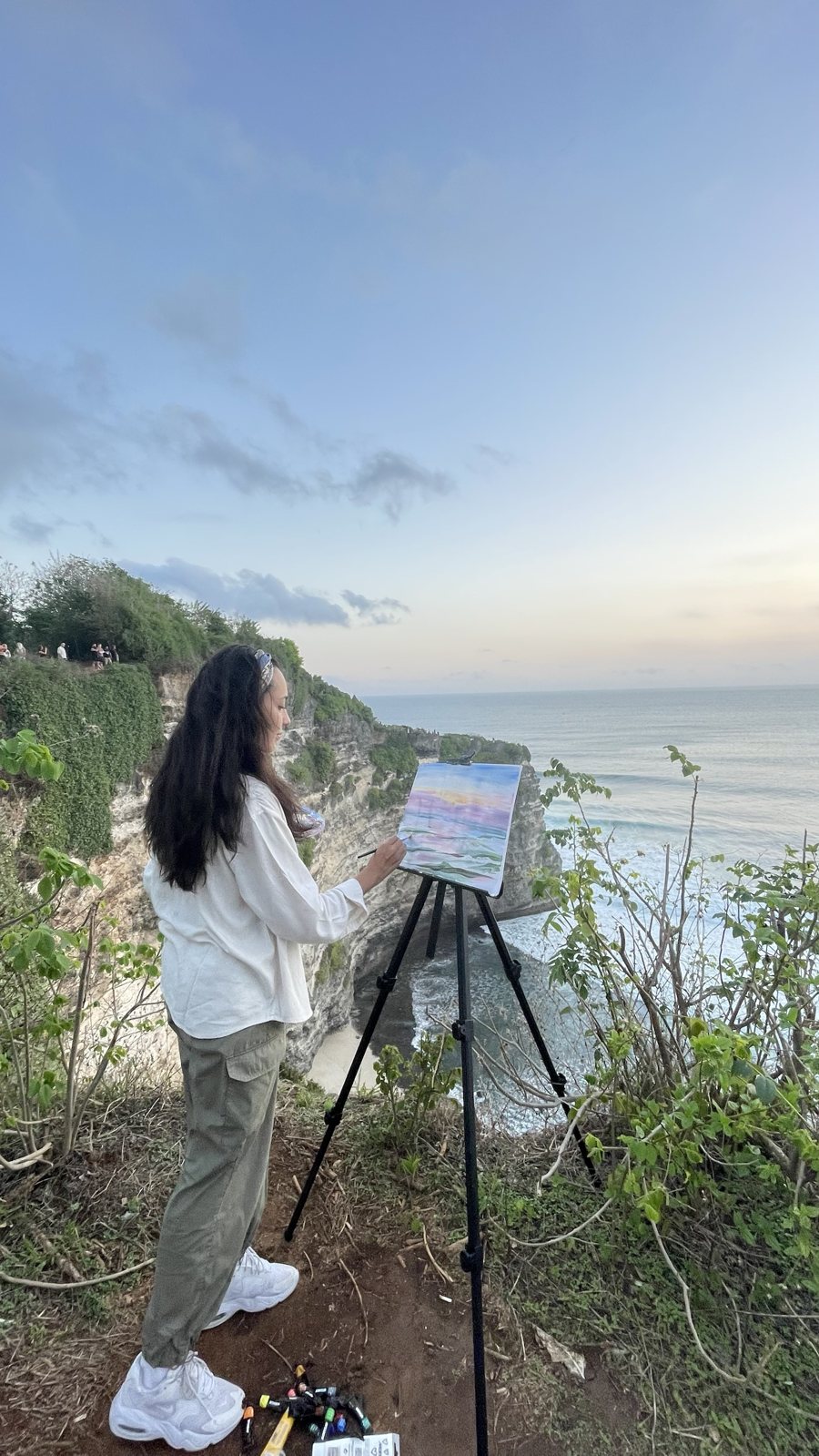 Aliya painting at an easel on a cliff above the ocean at sunset