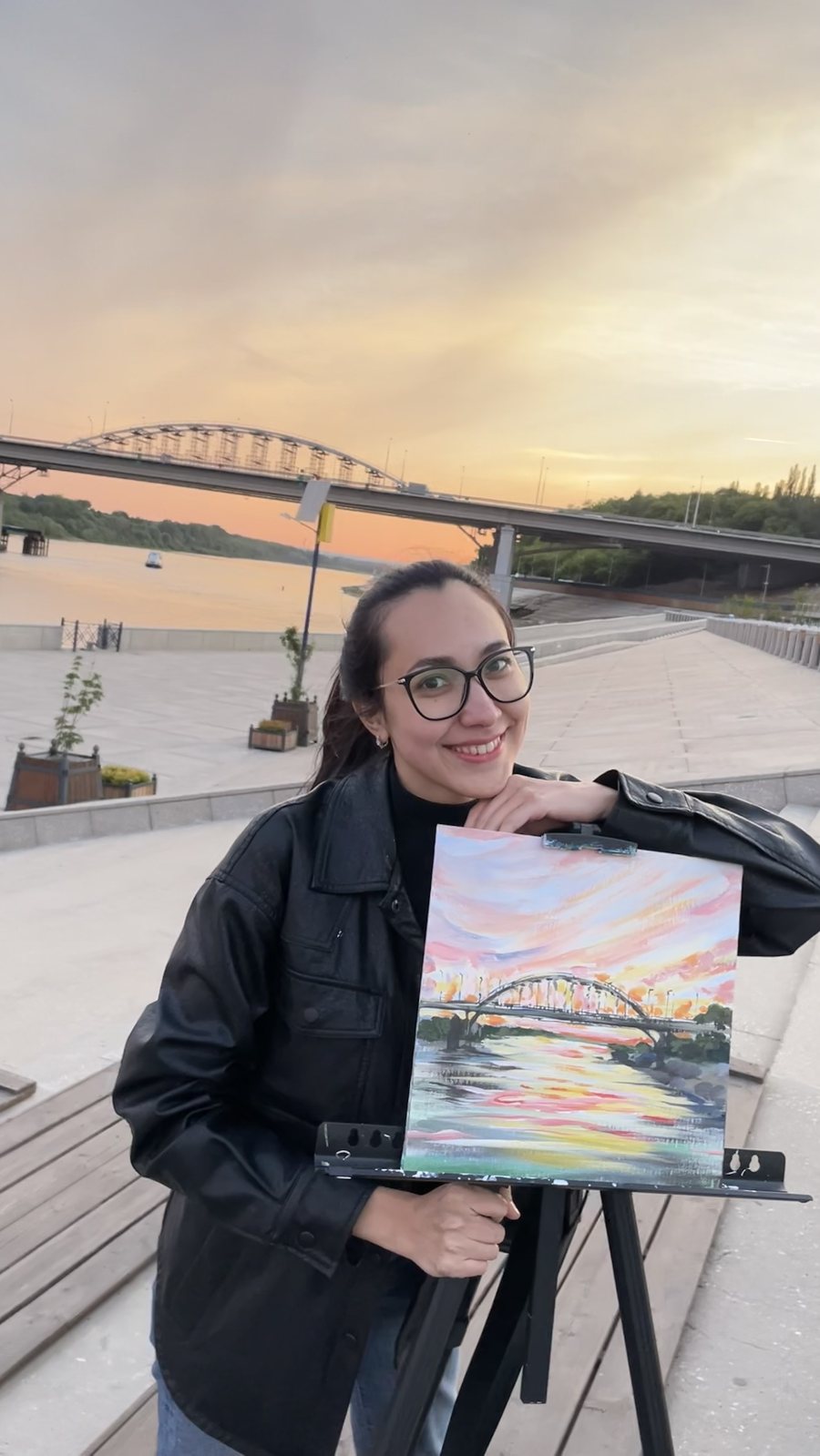 Aliya smiling beside her finished painting of the bridge at sunset