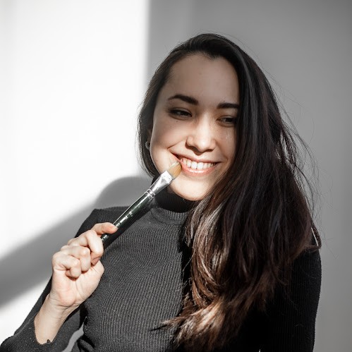 Aliya smiling in warm sunlight, holding a paintbrush to her lips